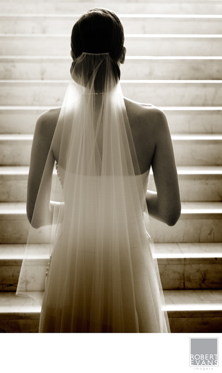 A bride is photographed from behind as light filters through window blinds and her veil.