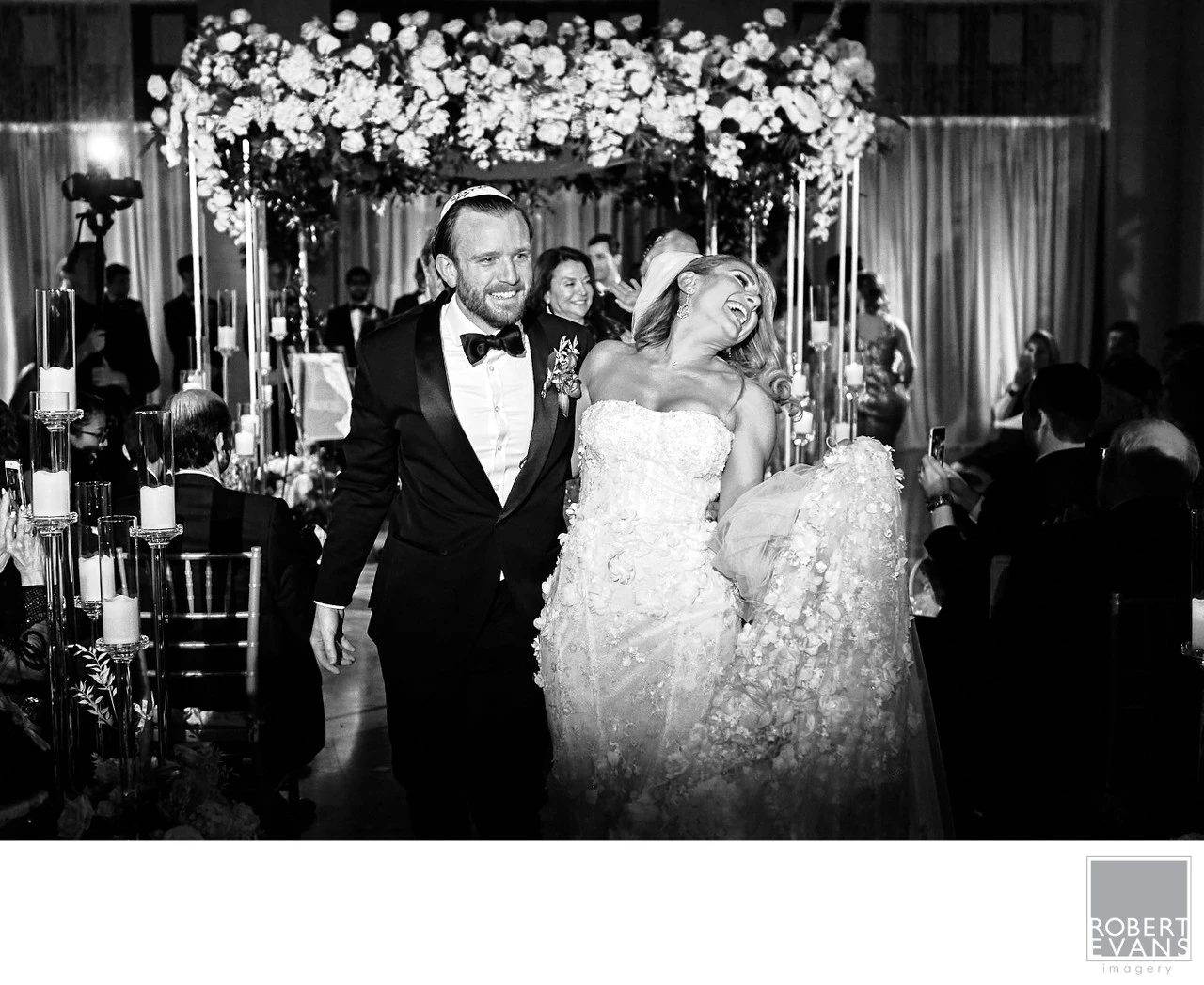A smiling bride and groom stand together beneath elaborate floral reception decorations in black and white.
