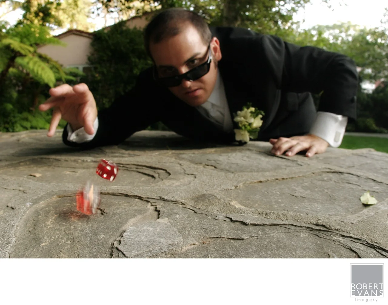 A man in formal clothing leans low over a stone surface as he rolls red dice.