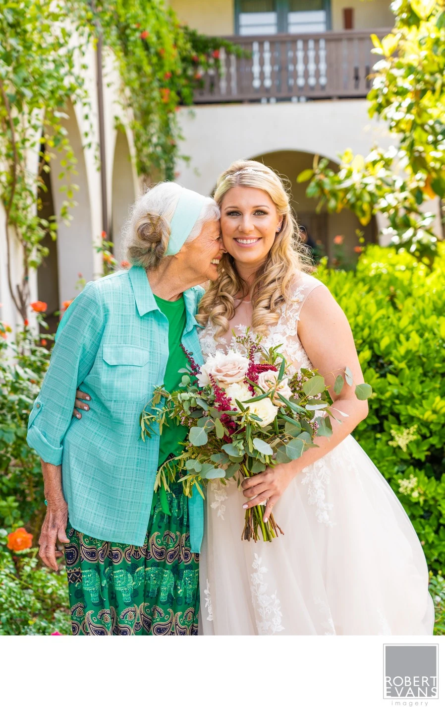 A bride holding flowers stands beside an older woman on a sunlit garden path.