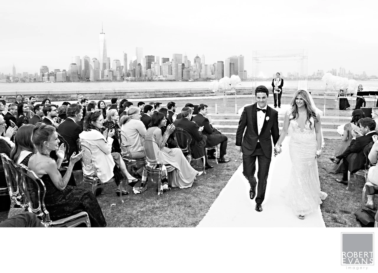 A bride and groom walk past seated guests with the New York skyline across the water in black and white.