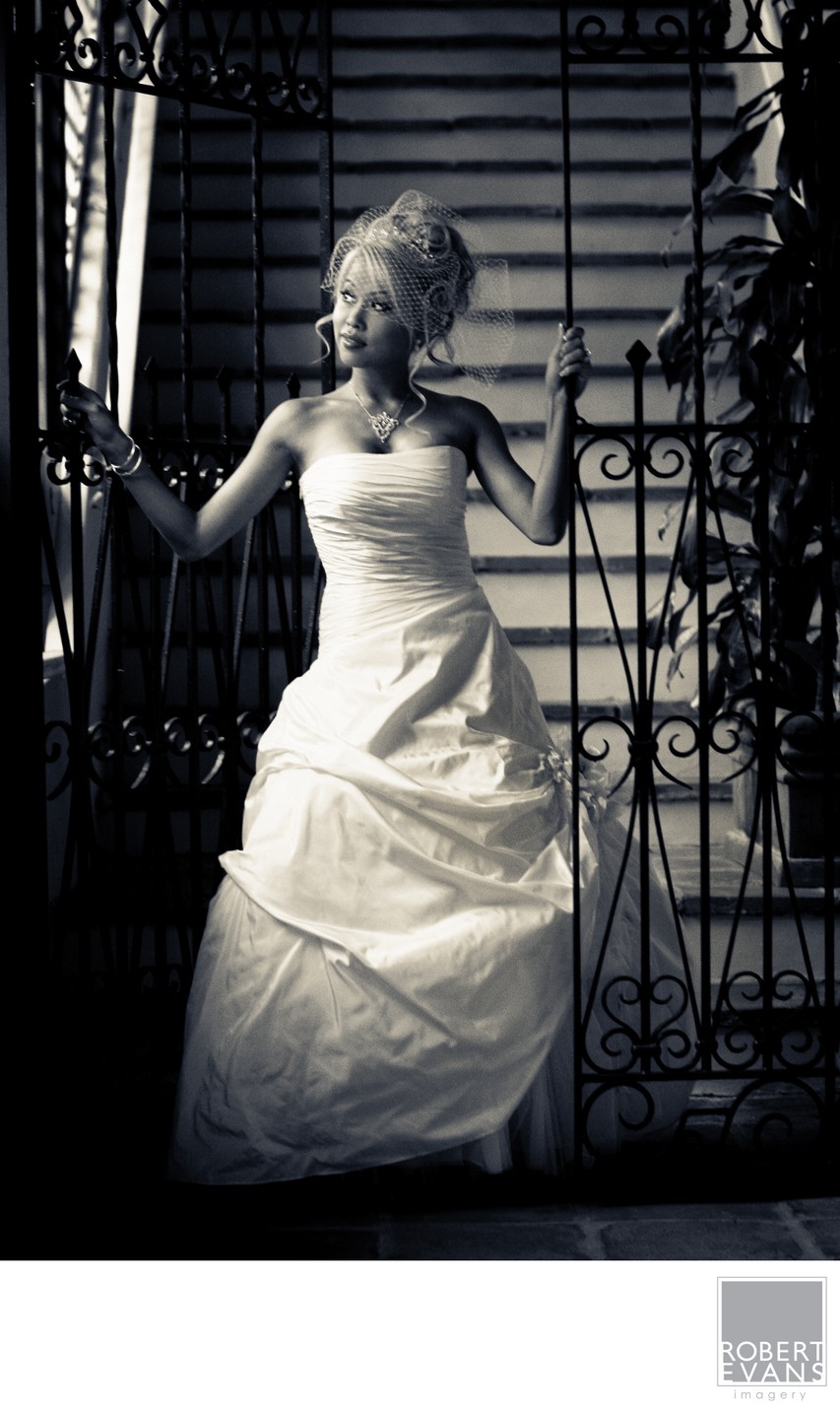 A bride stands against an ornate iron gate in a dramatically lit monochrome portrait.