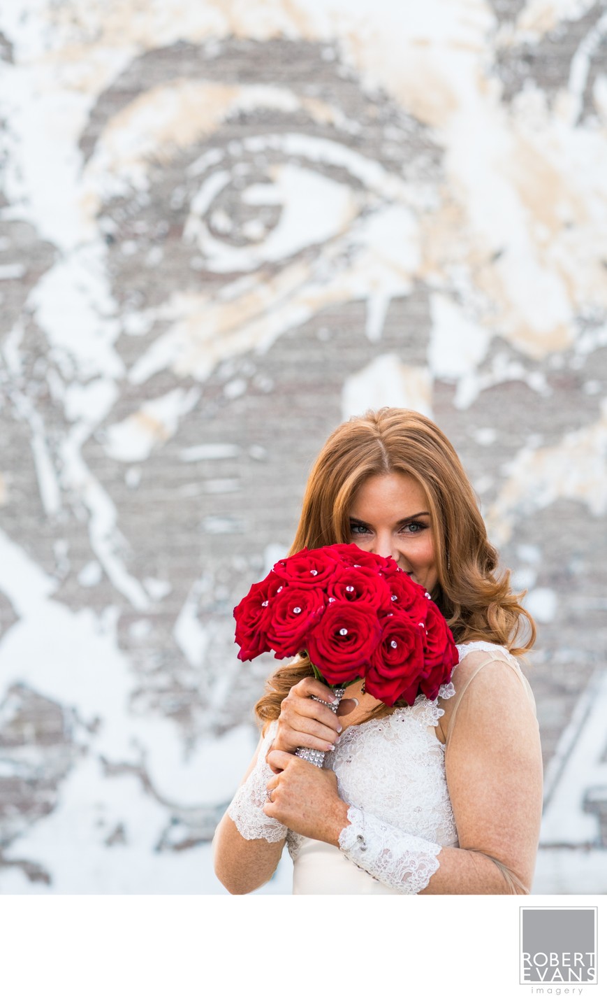 A bride holds red roses in front of her face with a large mural behind her.
