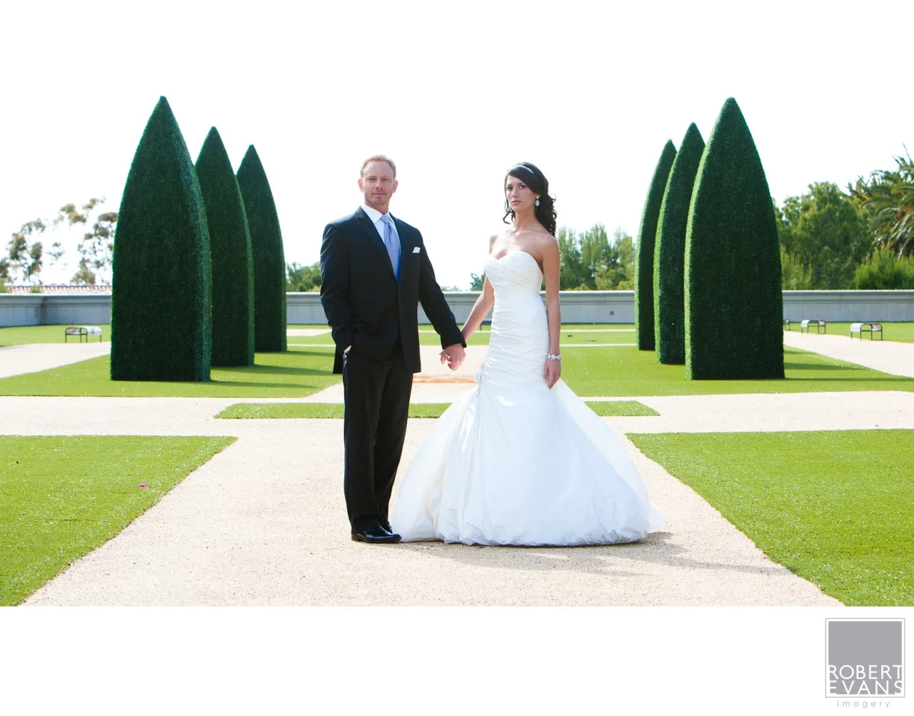 A bride and groom hold hands on a path bordered by sculpted green hedges.