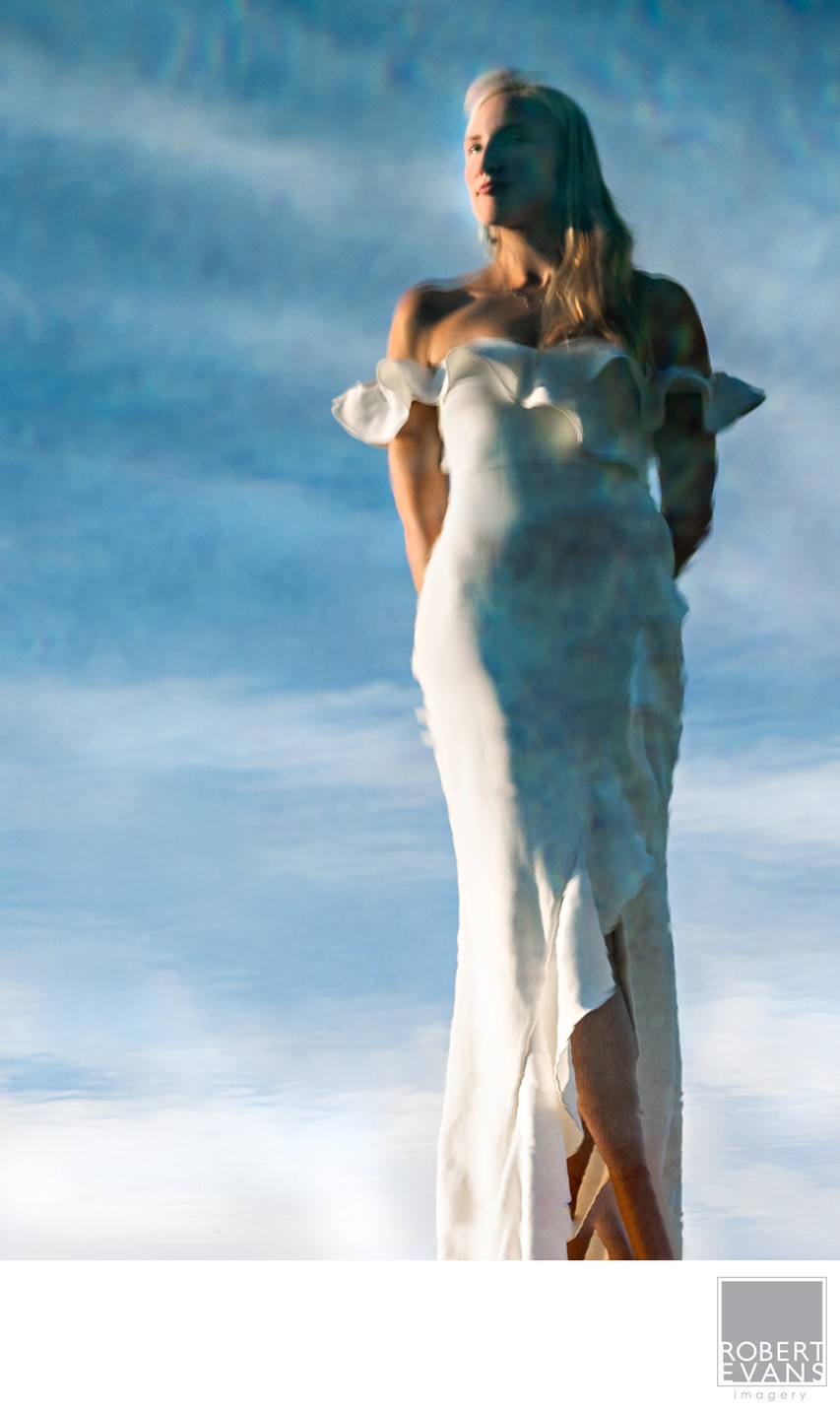 A bride in a fitted gown stands against a clouded blue sky in directional light.