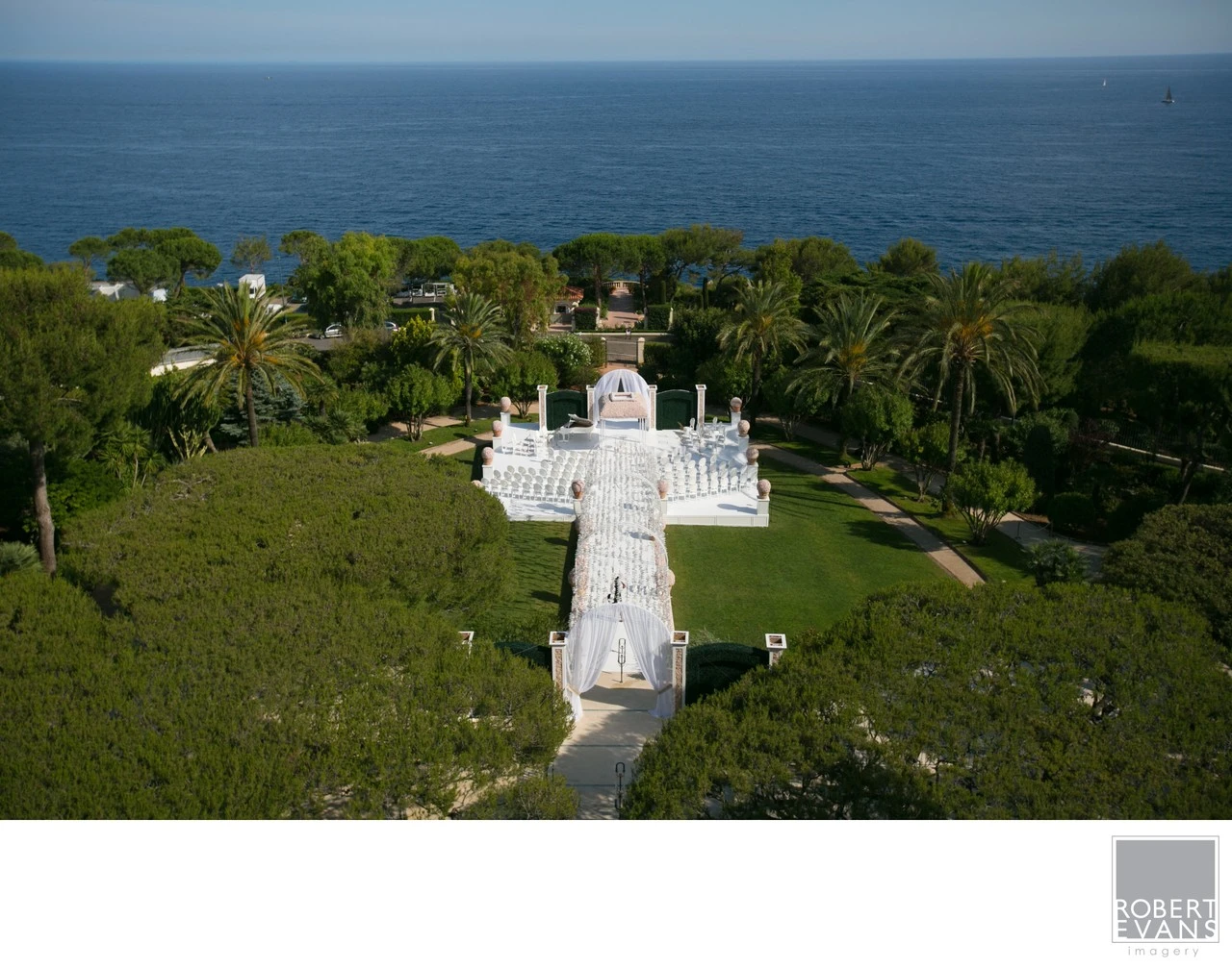 Rows of white chairs and a ceremony aisle overlook a broad blue ocean from a garden.