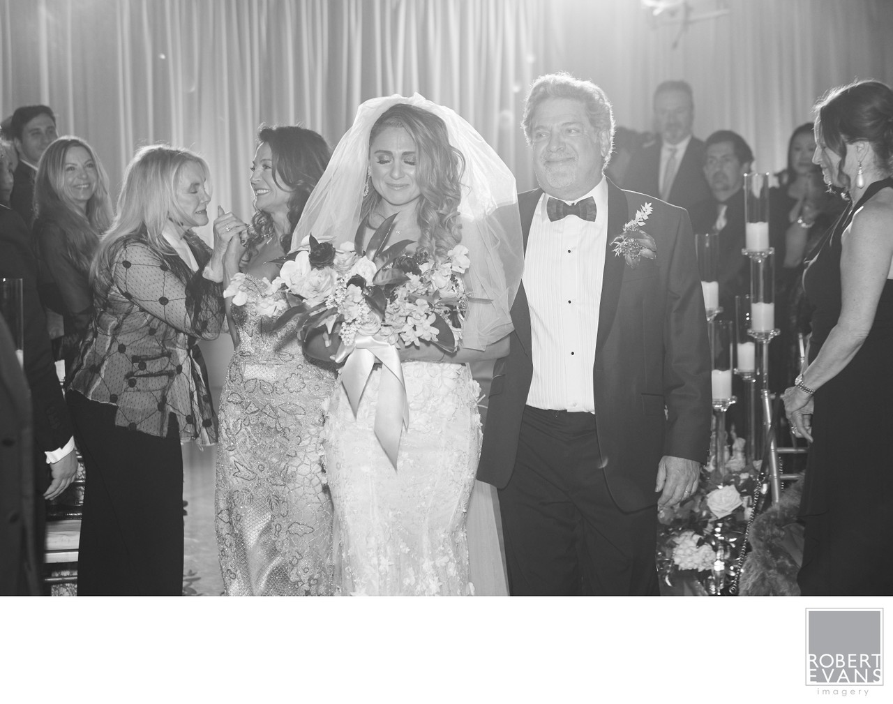 A bride walks beside her father as wedding guests watch in black and white.