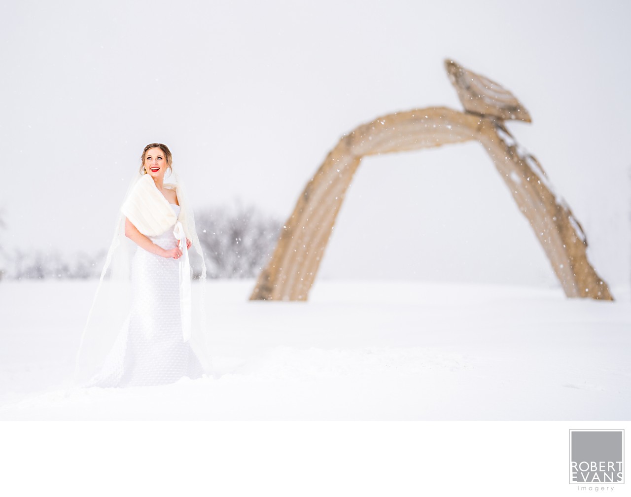 A bride stands on snow near a large sculptural wooden arch.