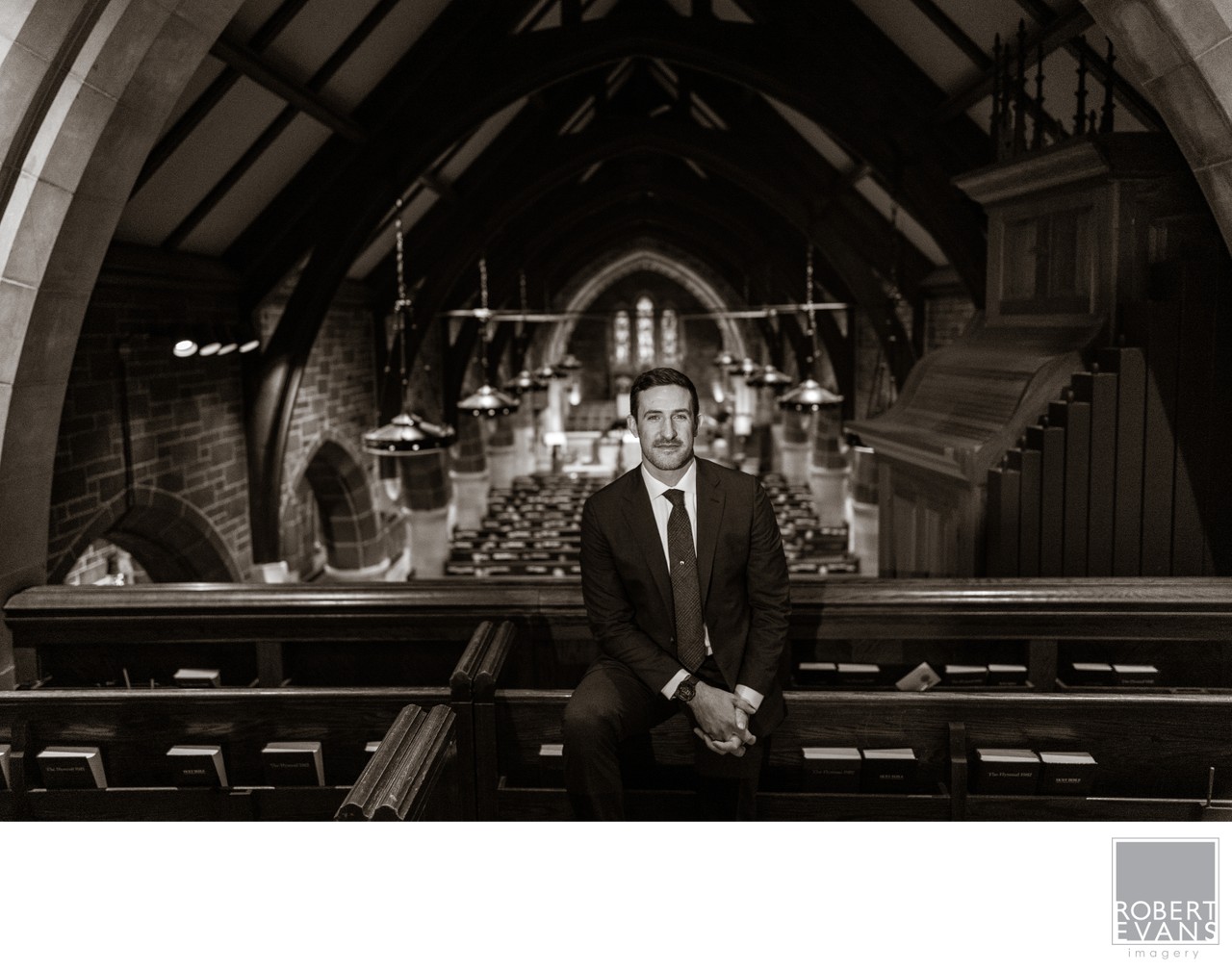 A groom stands between wooden church pews beneath a vaulted ceiling in black and white.