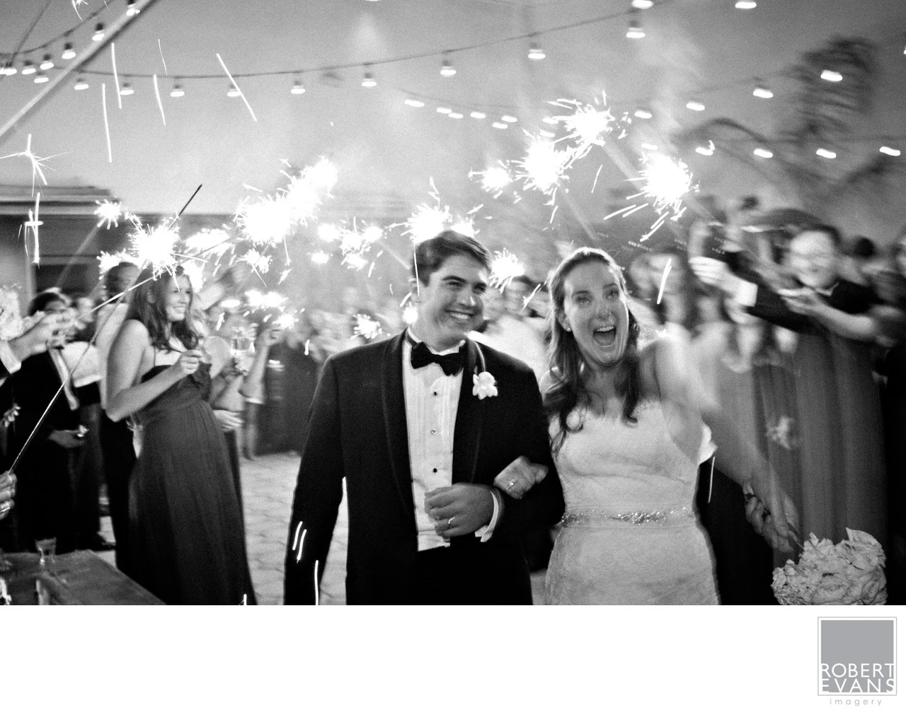 A bride and groom smile beneath sparklers held by wedding guests in black and white.