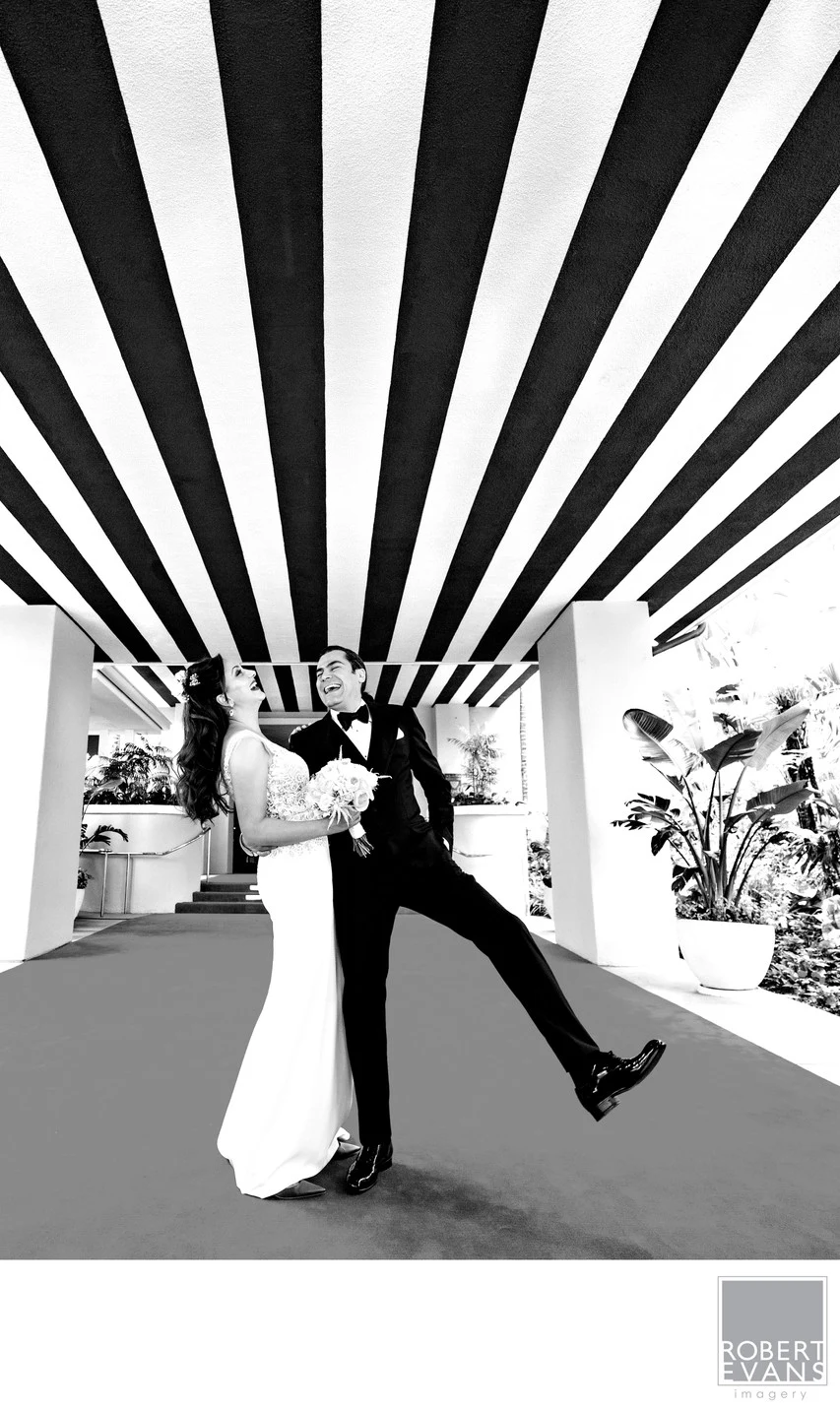 A bride and groom strike a playful pose beneath bold overhead beams in black and white.