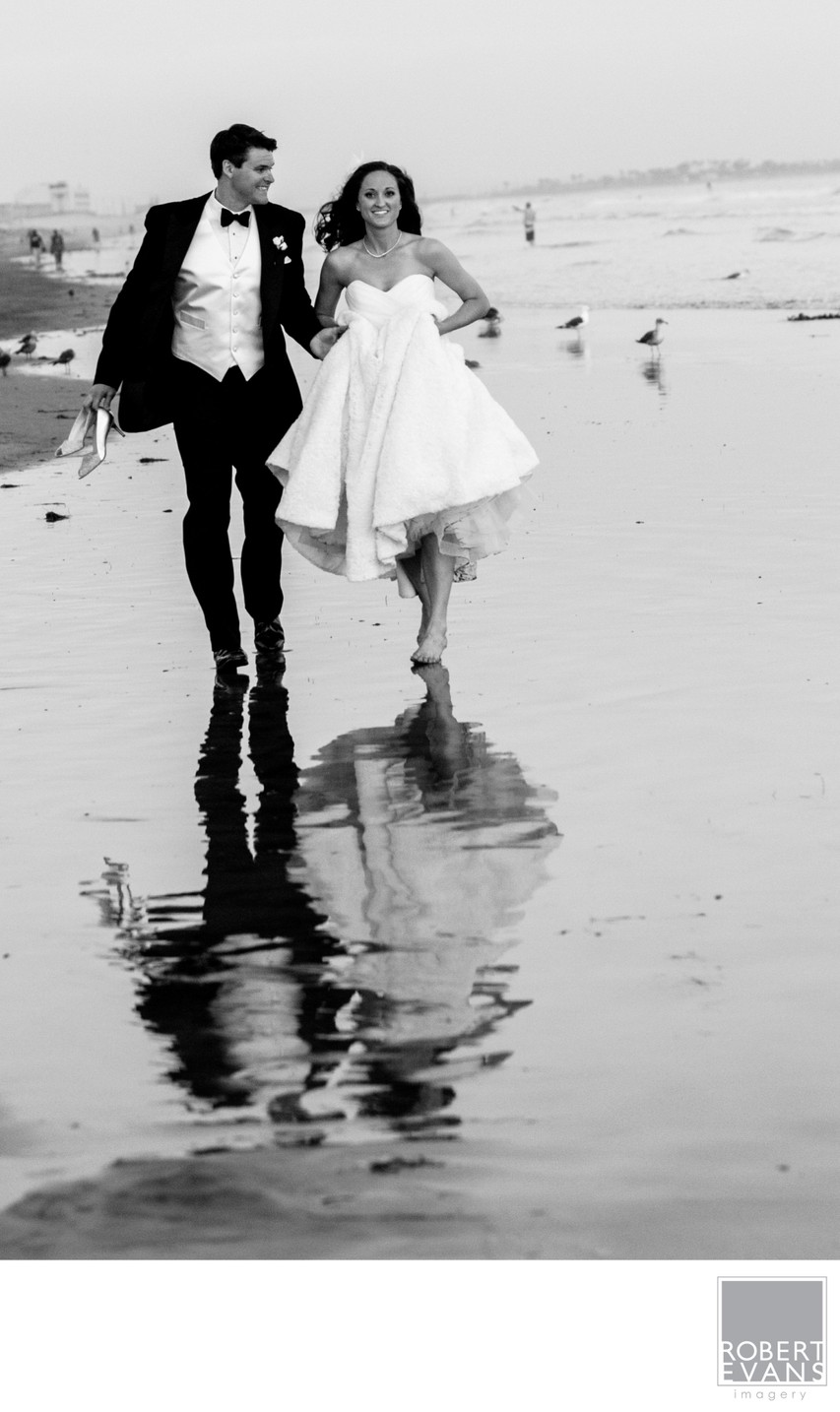 A bride and groom walk barefoot along wet sand with their reflections beneath them.