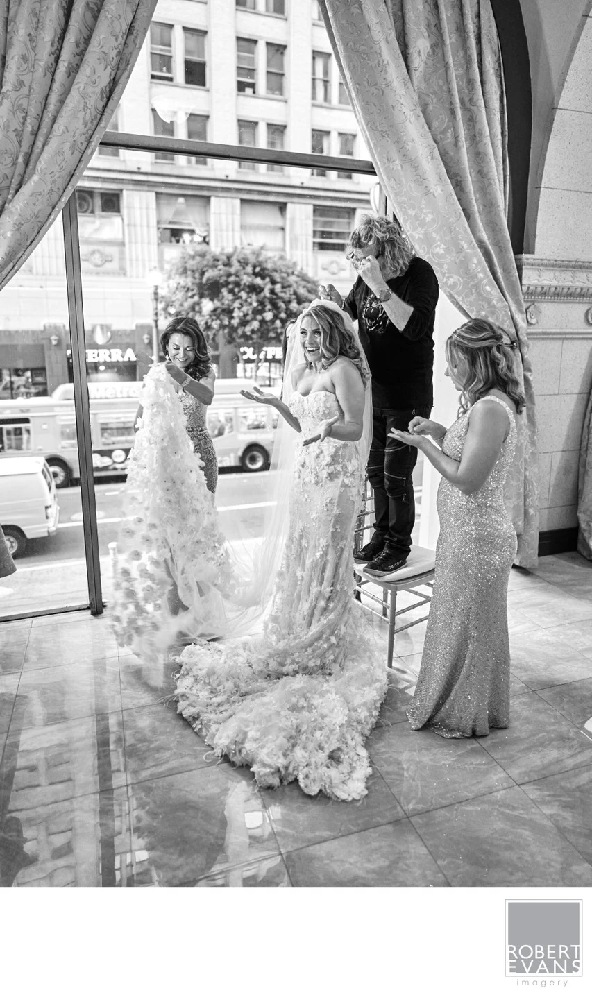 People lift an ornate wedding gown near tall windows during preparations in black and white.