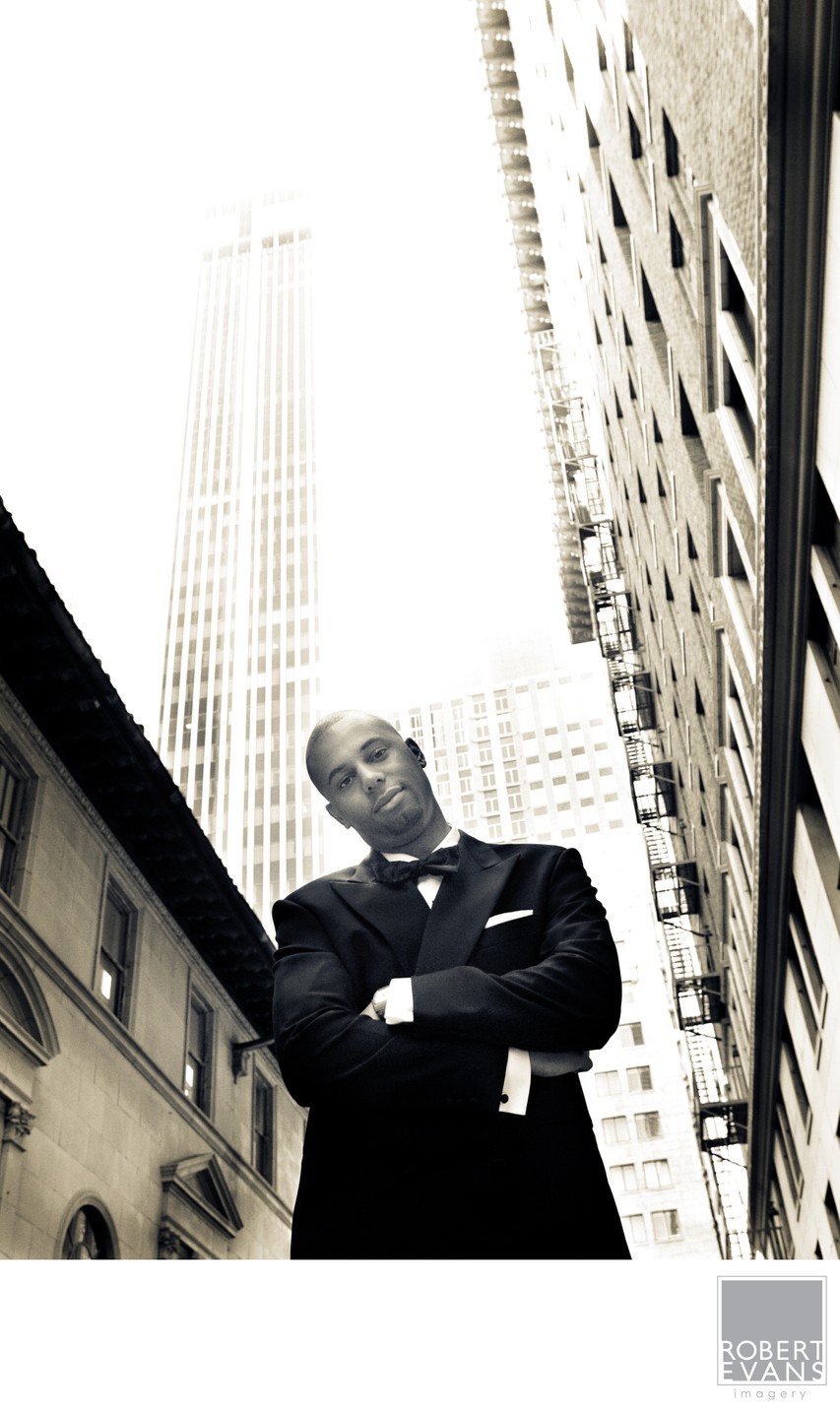 A groom stands with folded arms between tall buildings in a monochrome urban portrait.