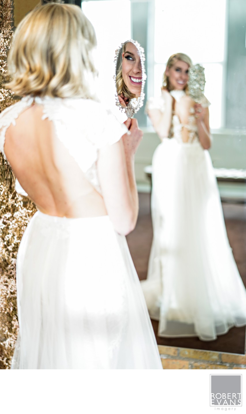 A bride in a white gown faces a mirror that reflects her smile and dress.