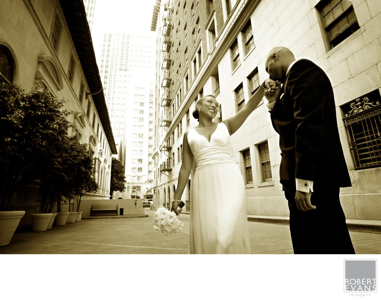 A groom kisses a bride’s raised hand between tall buildings in a warm monochrome portrait.
