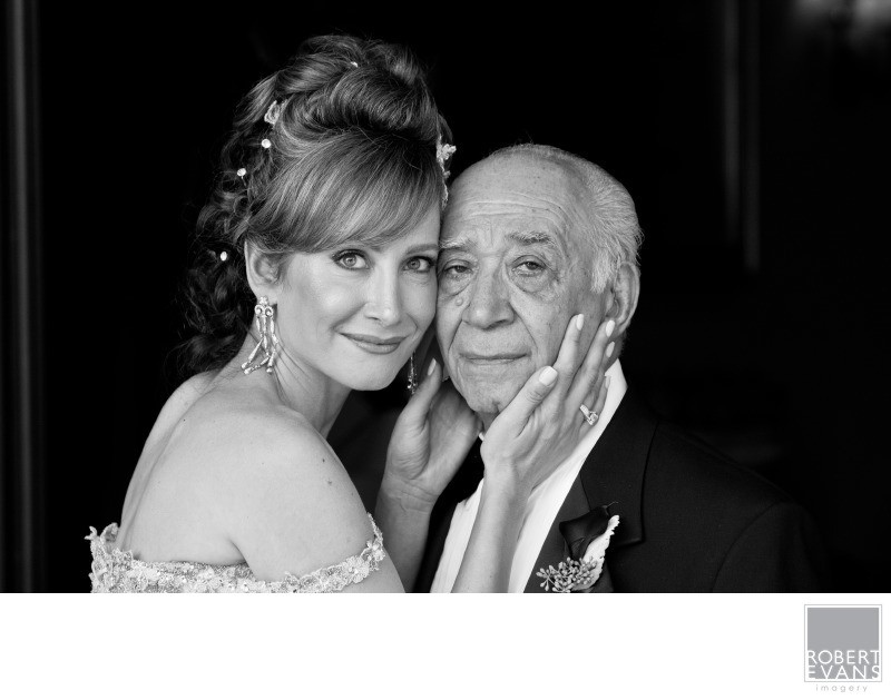 A bride presses her cheek against an older man and gently holds his face in black and white.