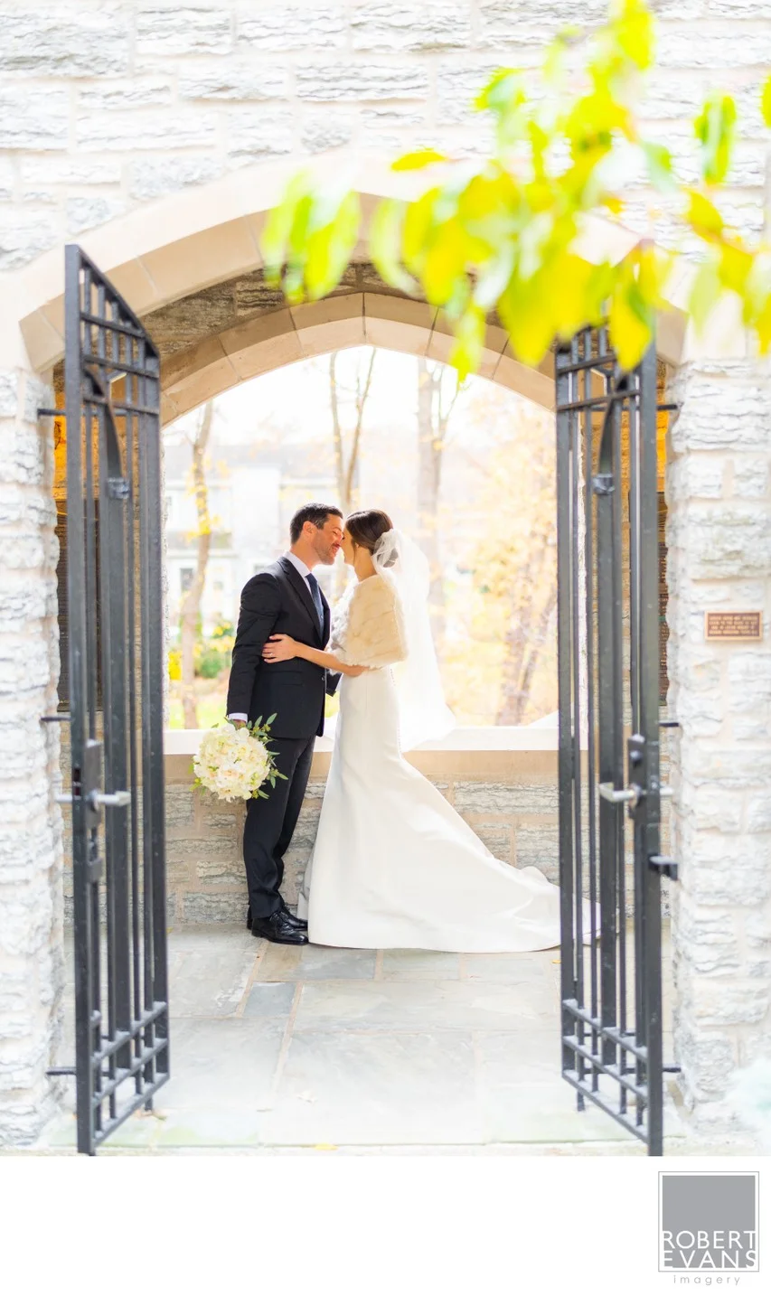 A bride and groom embrace beneath a stone arch through open iron gates.