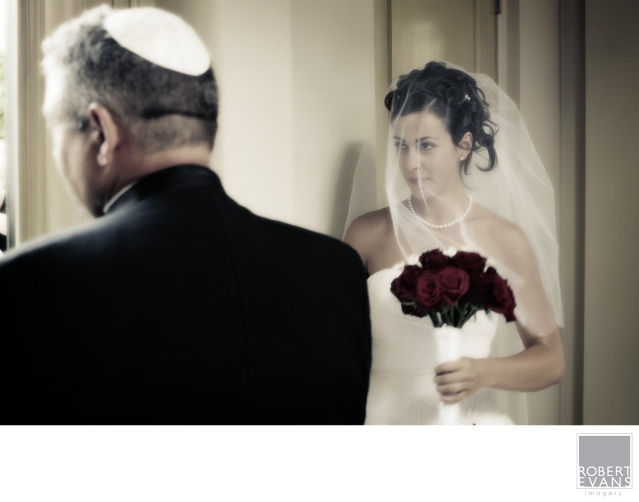 A bride holding dark red roses faces an older man wearing a kippah during the ceremony.