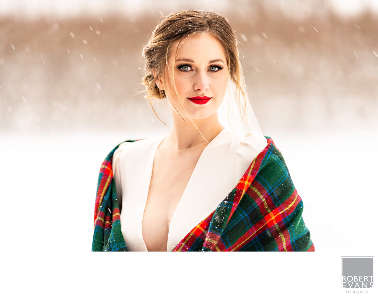 A bride wearing a colorful tartan wrap looks toward the camera as snow falls.