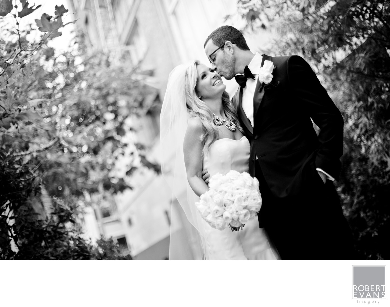 A bride and groom kiss outdoors beneath leafy branches in black and white.