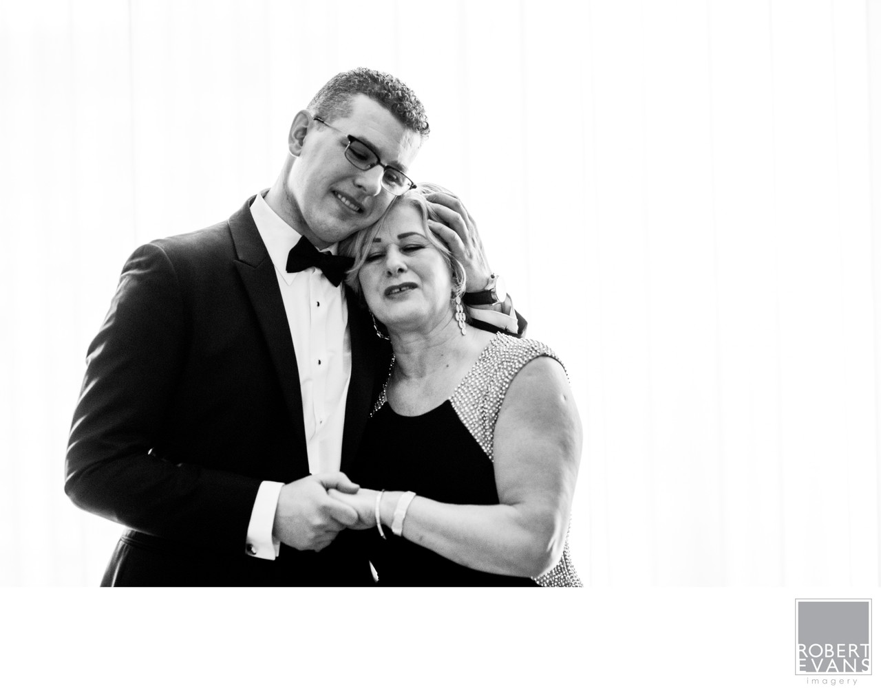 A groom holds an older woman close as they dance in black and white.