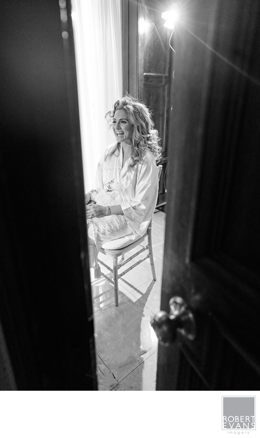 A bride sits beside a tall window seen through a partly open doorway in black and white.