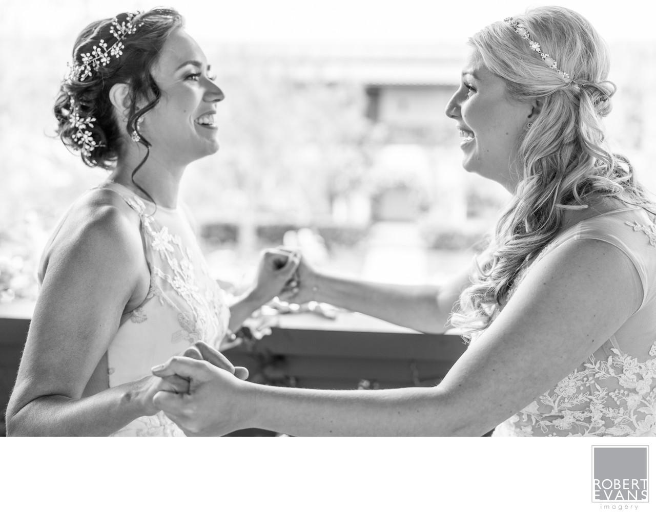 Two women in wedding attire hold hands and laugh together in black and white.