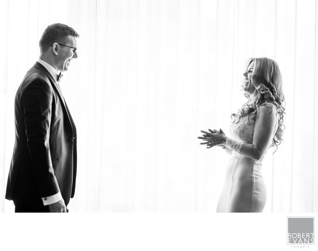 A groom and bride face each other against bright curtains in black and white.