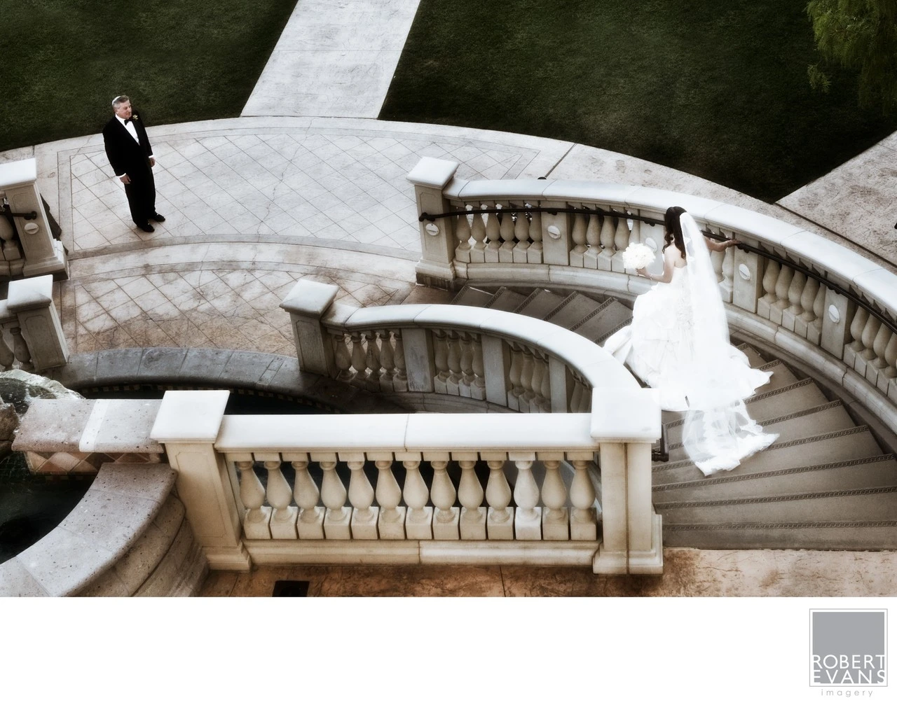 A groom waits above while a bride moves down a sweeping outdoor stone staircase.