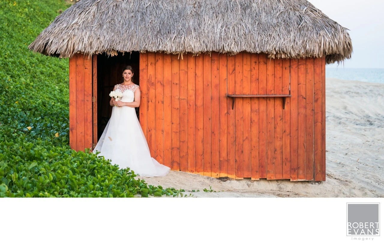 A bride in a white gown stands beside an orange wooden hut with a thatched roof.