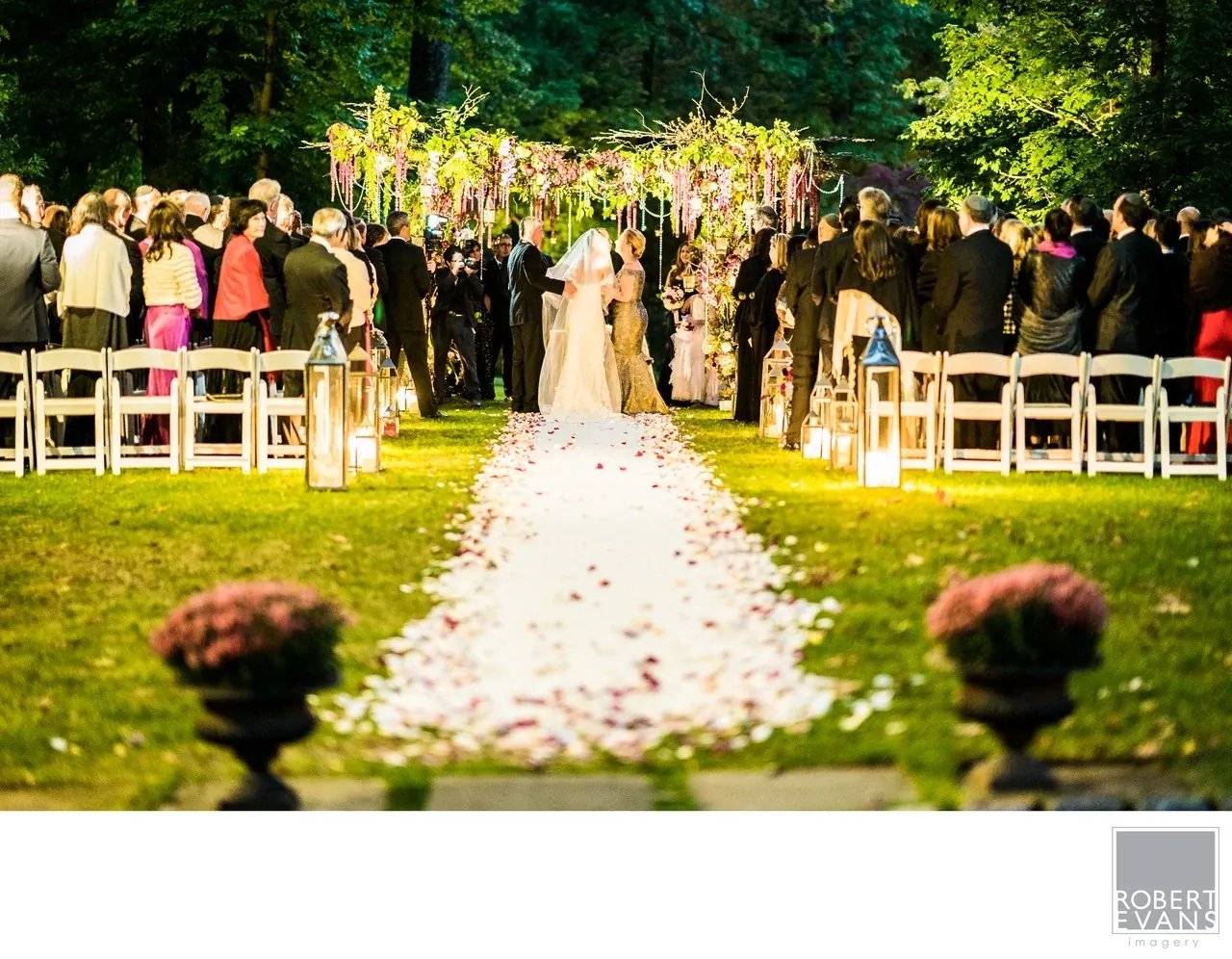 Guests stand beside a petal-covered aisle during an outdoor ceremony beneath greenery and flowers.