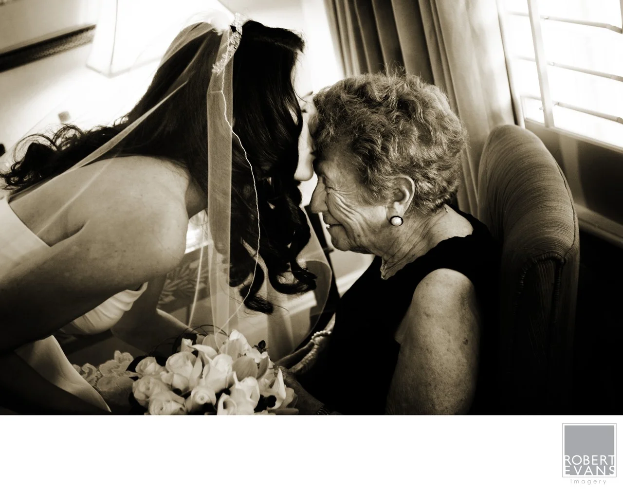 A bride leans close to an older woman seated indoors in a warmly toned monochrome photograph.