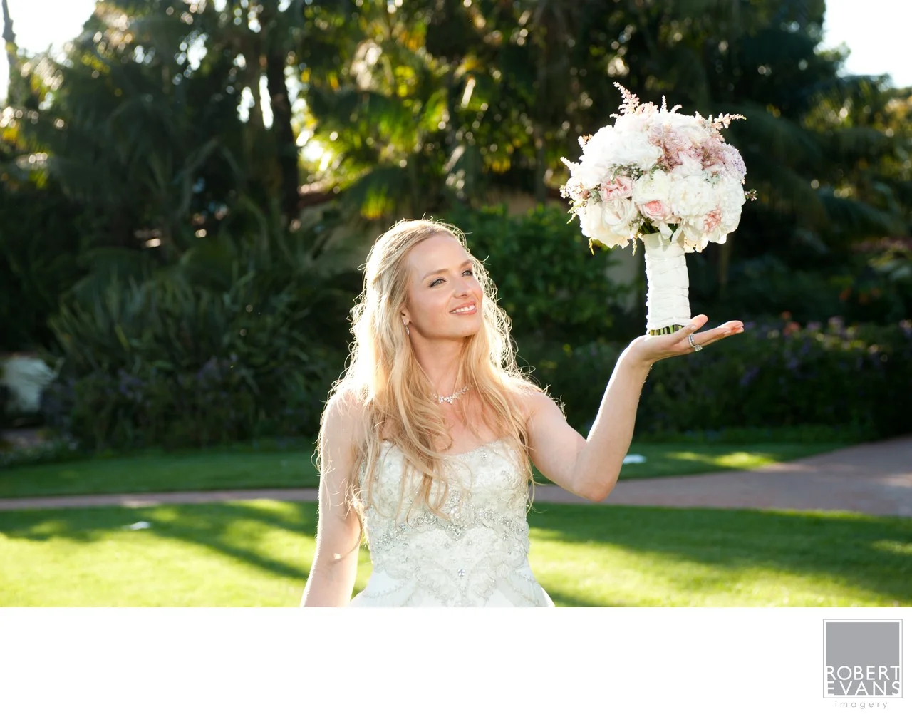 A smiling bride balances a pale bouquet above her open hand outdoors.