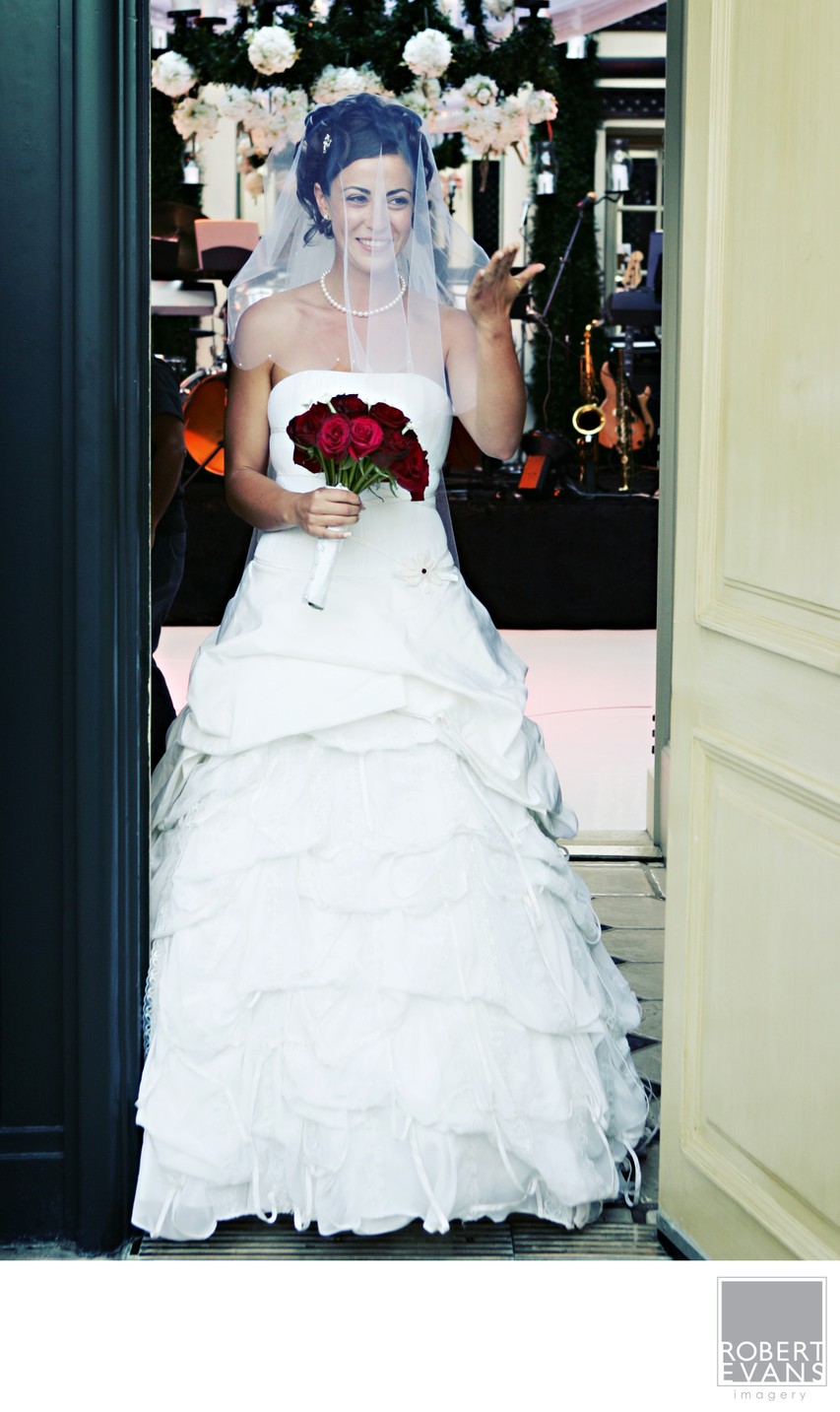 A bride holding a red rose bouquet stands in a doorway during wedding preparations.
