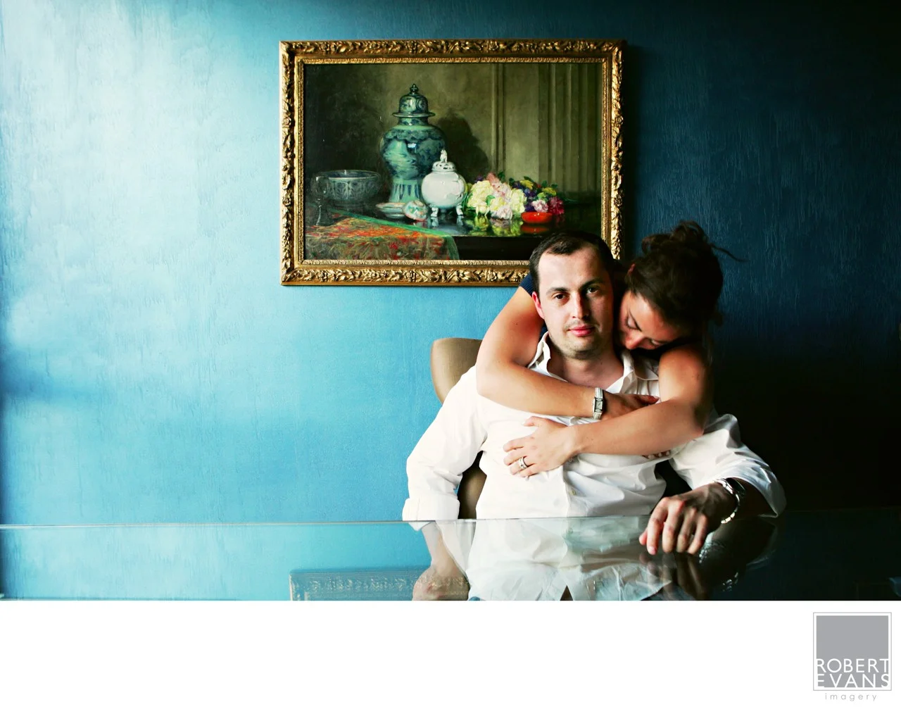 A bride embraces her groom from behind beneath a framed still-life painting on a blue wall.
