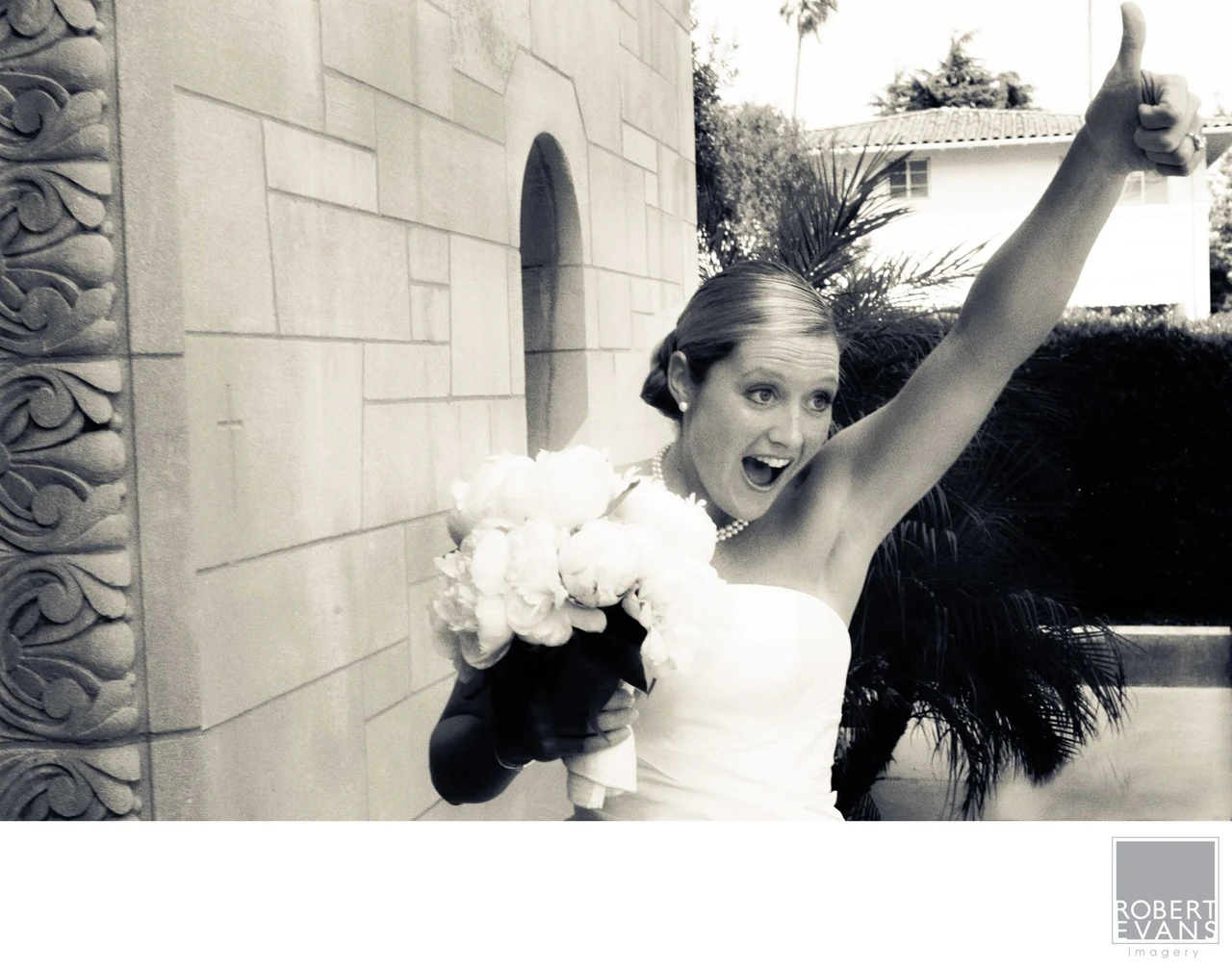 A bride lifts her arm high and smiles beside a stone building in black and white.
