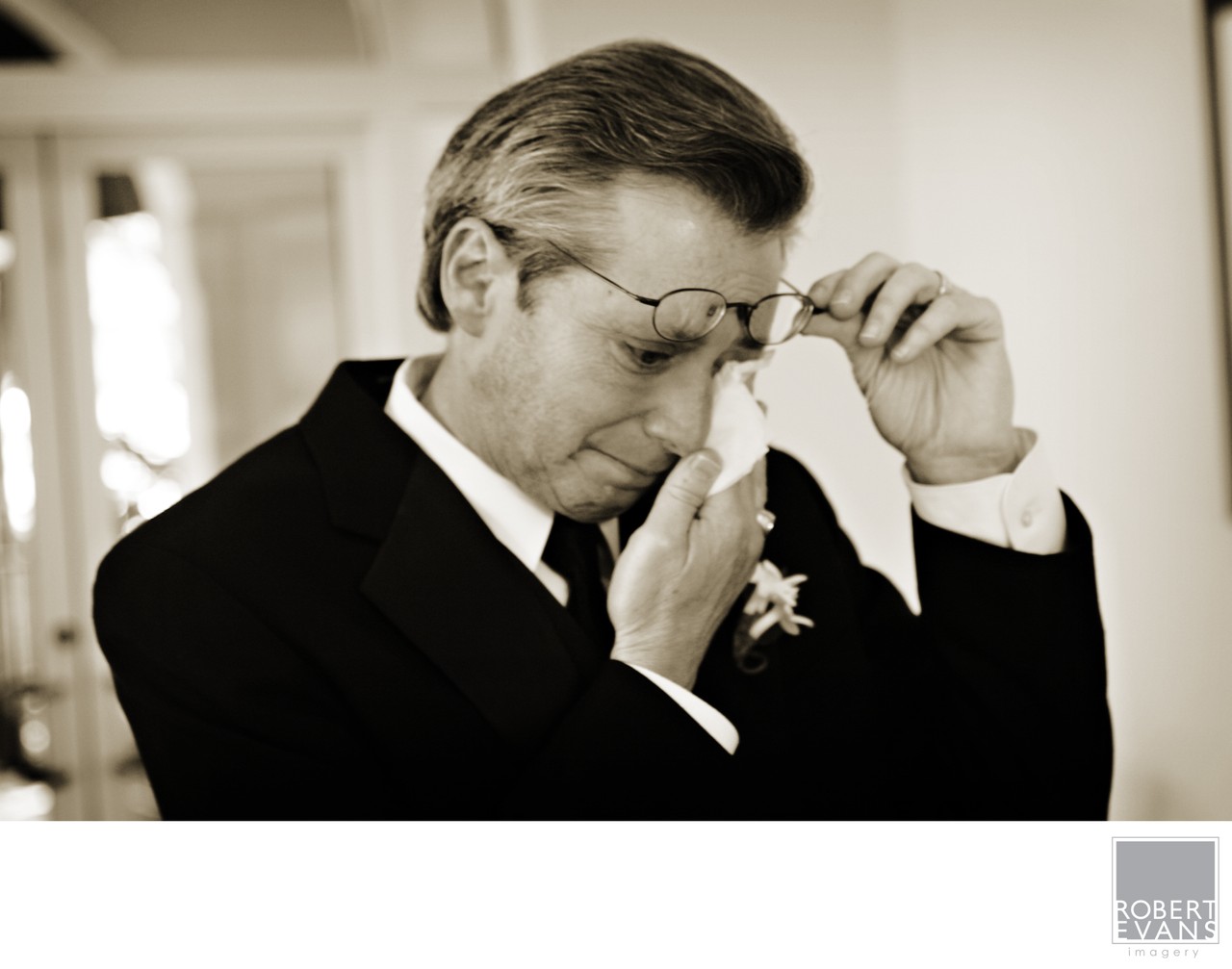 A man in formal clothing lifts his glasses and wipes his eye in a close monochrome portrait.