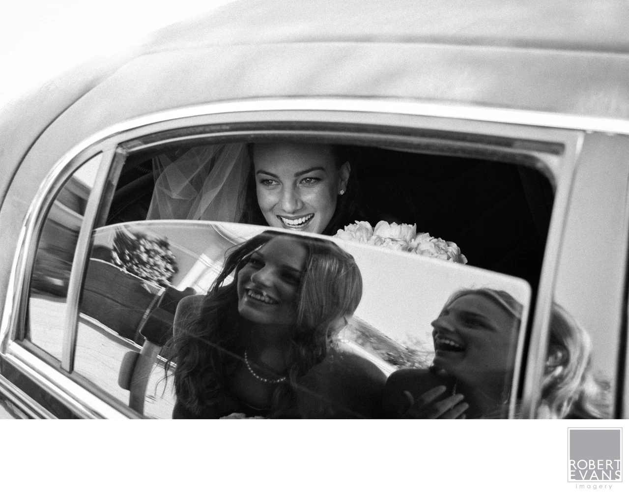 A smiling bride looks through a partly lowered car window reflecting women standing outside.