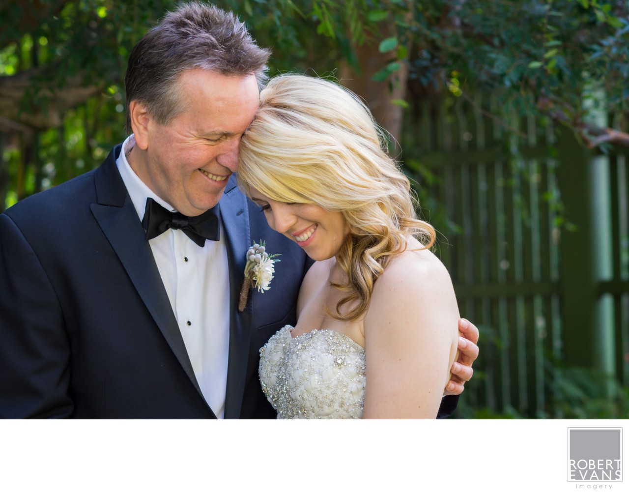 A bride rests her head against her groom’s chest in warm outdoor light.