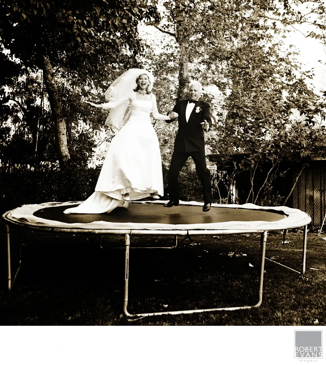 A bride and groom bounce on a trampoline beneath leafy trees in black and white.