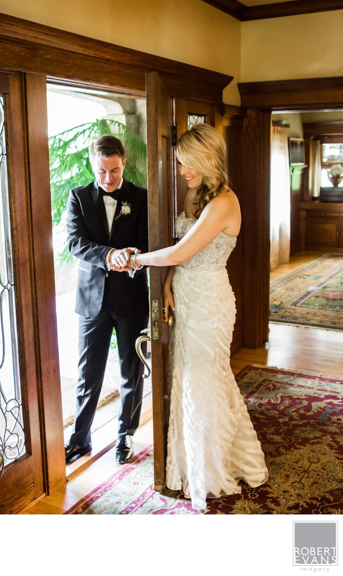 A bride and groom hold hands beside a sunlit doorway during a private wedding-day moment.