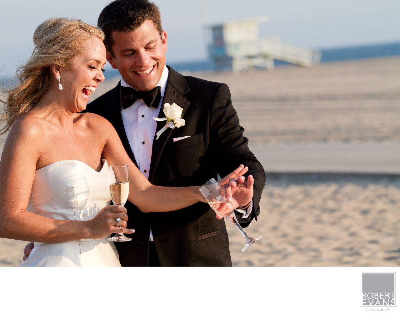 A bride and groom hold drinks and smile together on a sandy beach.
