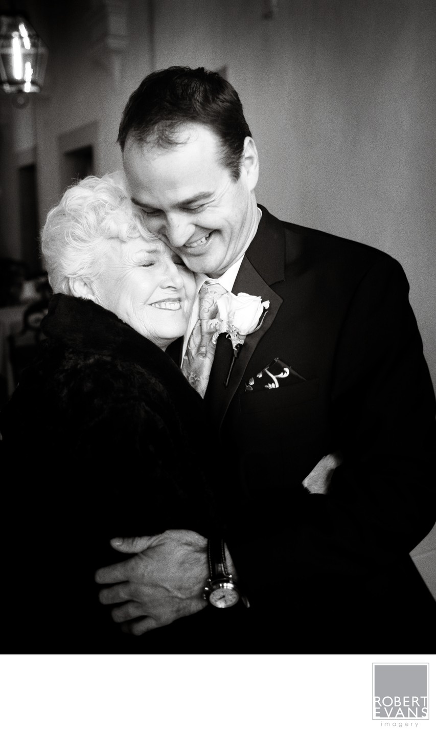 A groom embraces an older woman with his eyes lowered in black and white.