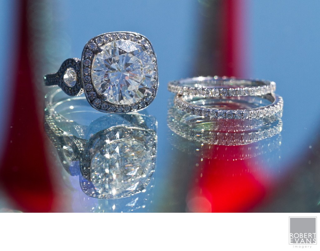 An engagement ring and wedding bands sparkle on a reflective surface against colored light.