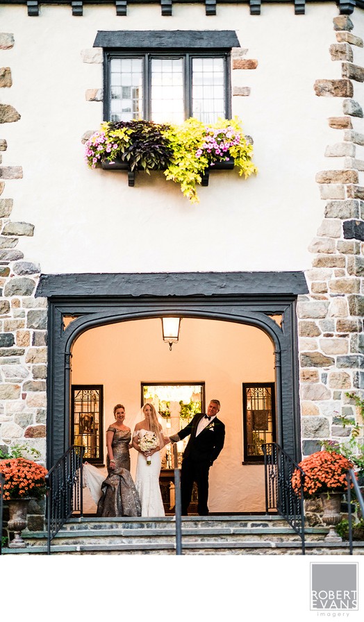 A bride and groom stand in an arched doorway beneath flower-filled windows.
