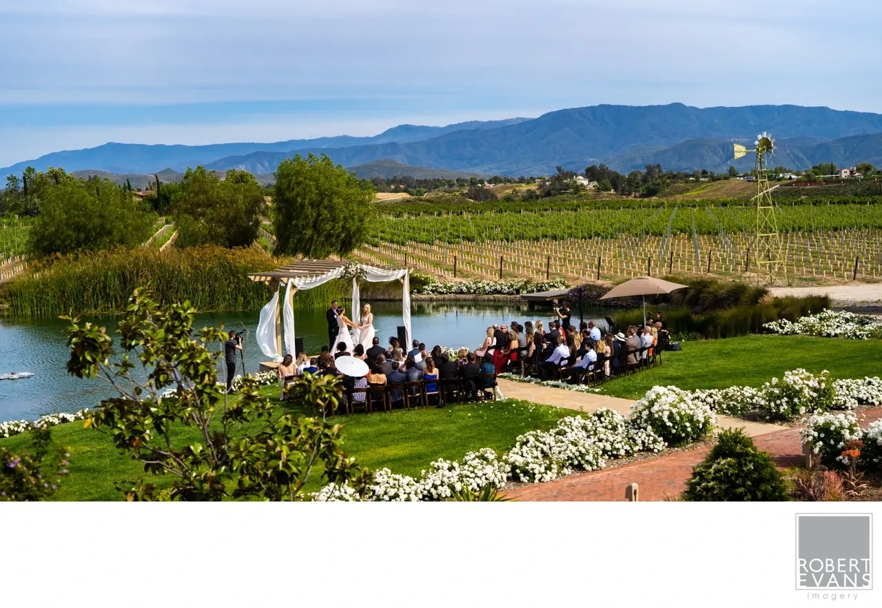 Guests attend an outdoor wedding beside water with vineyards and mountains beyond.