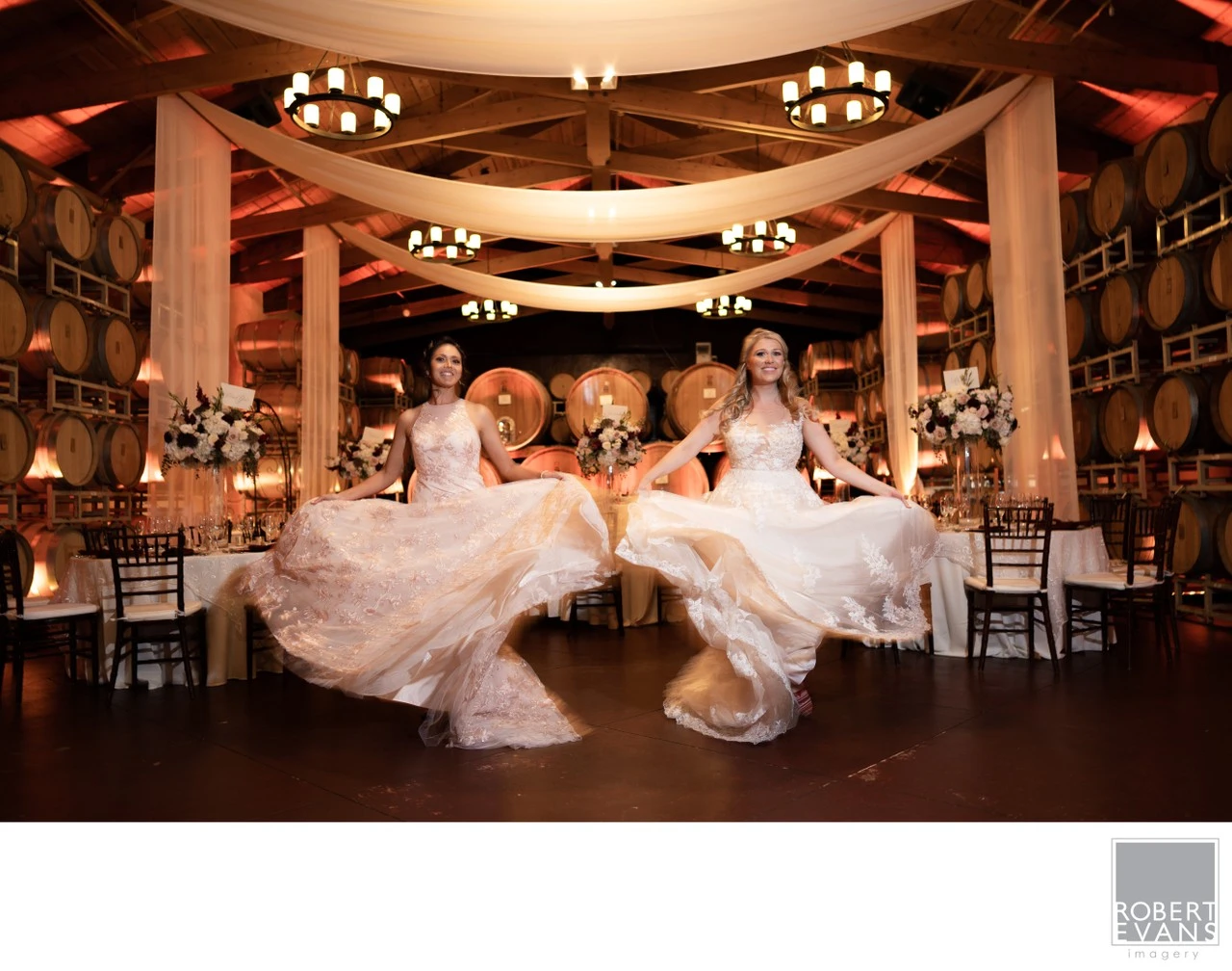 Two women twirl in wedding gowns beneath draped fabric and chandeliers at a reception.