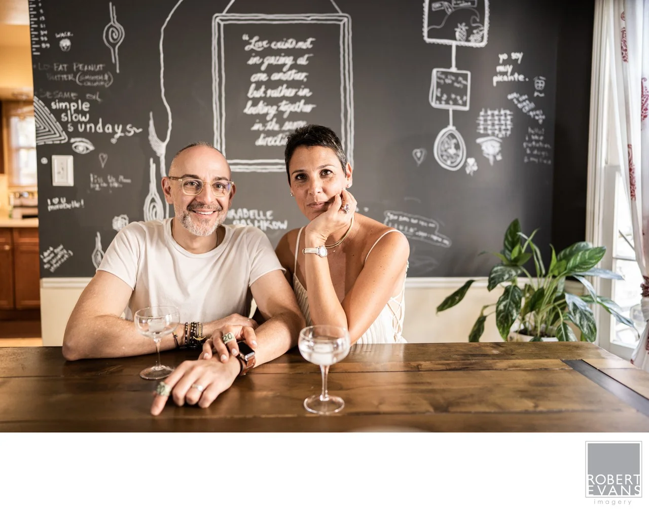 A smiling couple sits side by side at a wooden café table with wine glasses.