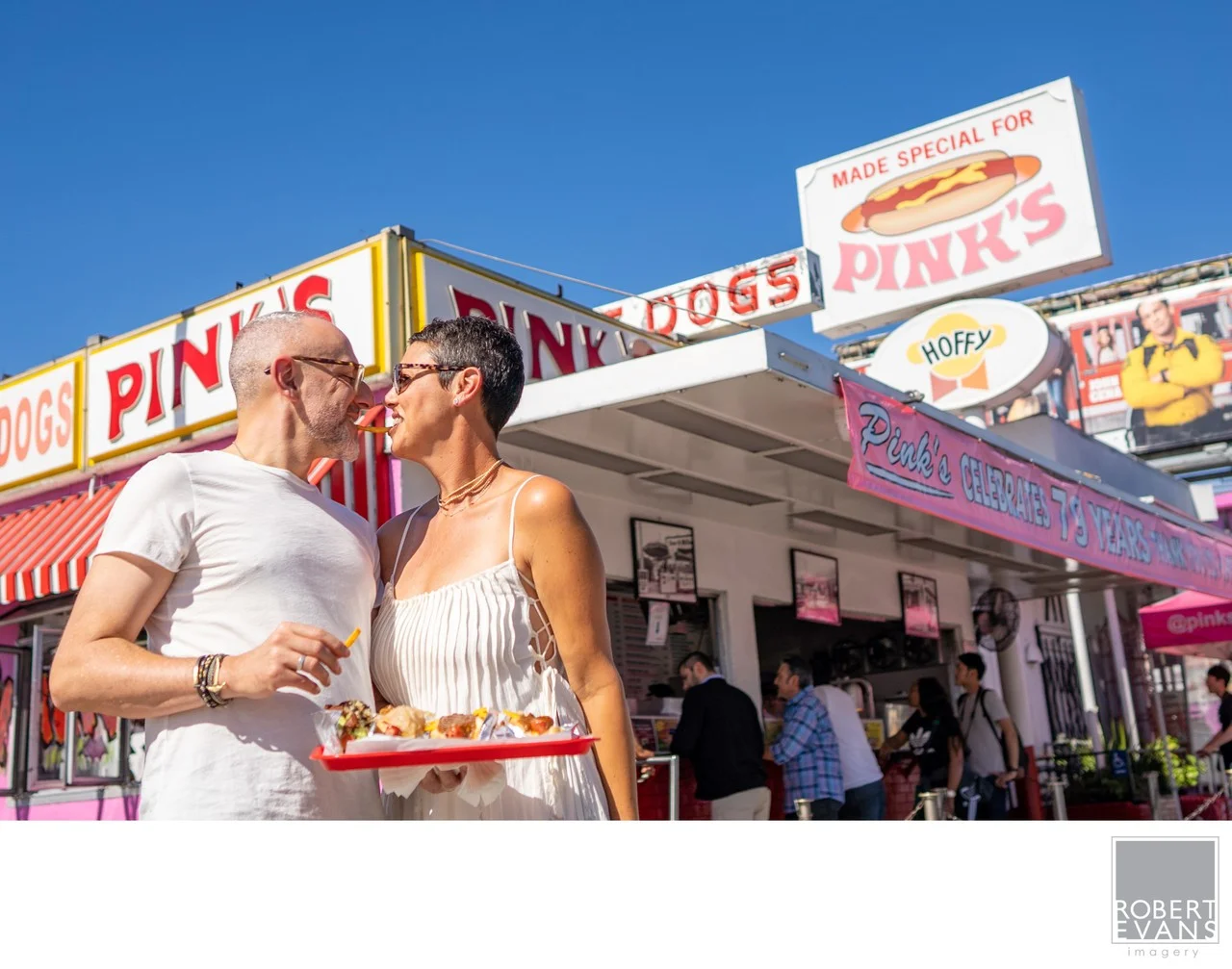 A couple holds a tray of food outside a colorful hot dog stand.